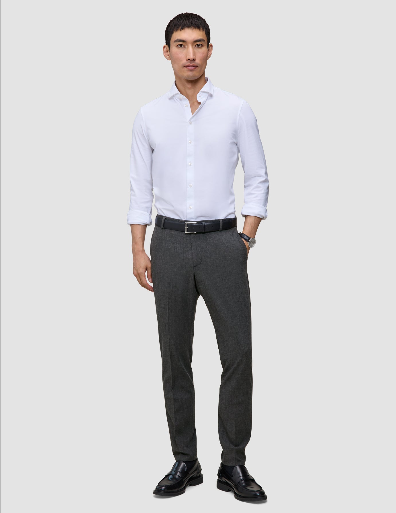 En mand iført en hvid skjorte med knapper, Motion Suit-bukser i Slim-pasform i farven Pepper Grey, et sort bælte og sorte loafers.